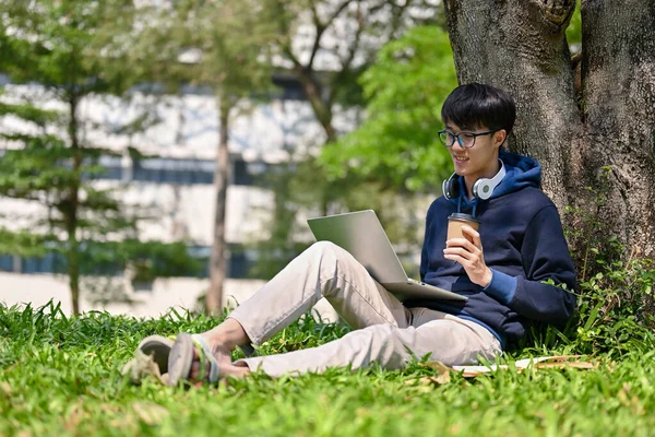 快乐的年轻亚洲大学生坐在校园里的一棵树下 一边用笔记本电脑管理功课 一边喝着咖啡 — 图库照片