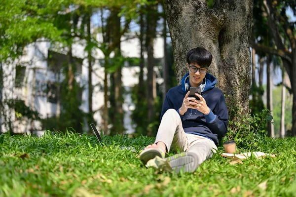 快乐的年轻亚洲大学生坐在校园里的一棵树下 一边用智能手机 一边玩手机游戏 — 图库照片