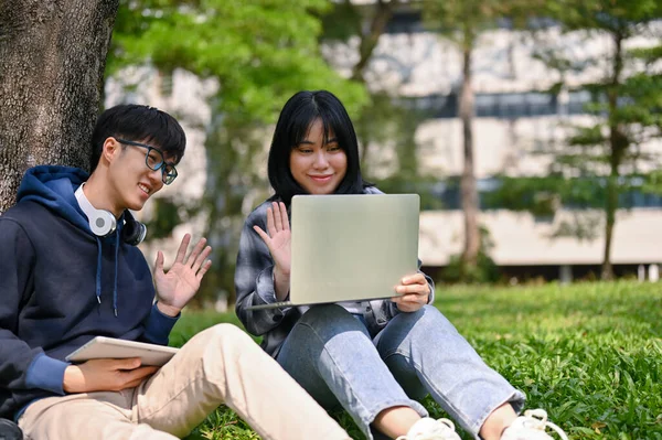 两名笑着 快乐的年轻亚洲大学生正坐在校园公园的草地上与老师举行在线会议或在线课程 — 图库照片