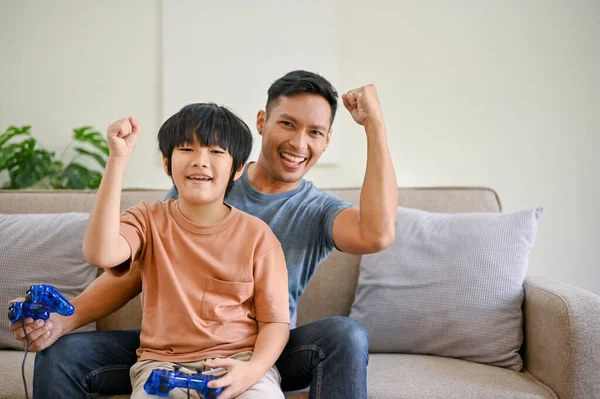 Joyful Asian dad and son with joysticks are playing video games and spending fun time at home together.