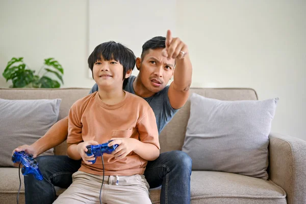 Excited Asian dad coaching his son to play video games with joysticks in the living room, spending fun family time together at home.
