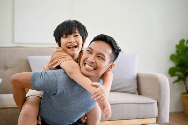 Joyful and happy Asian dad playing with his little son in the living room, piggyback, spending fun time together at home.