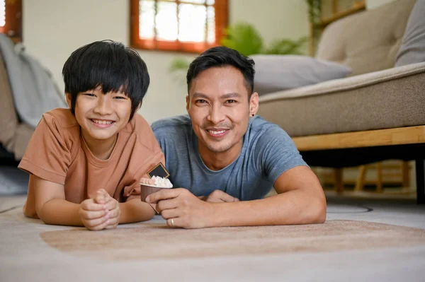 Happy and smiling Asian dad and son laying on the living room floor together, spending fun time on the weekend together.