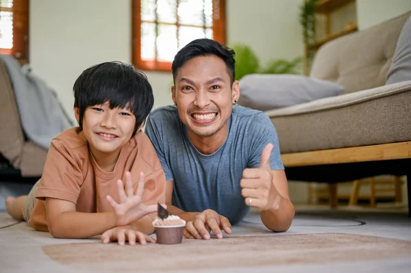 Happy and smiling Asian dad showing thumb up while laying on the living room floor with his son. happy family time concept