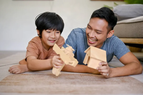 Happy Asian little boy playing a wooden figure model with his dad while laying on the living room floor together.