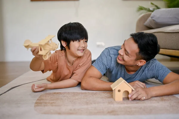 Playful and happy Asian little boy playing a wooden figure model with his dad while laying on the living room floor together. parenting and childhood concept