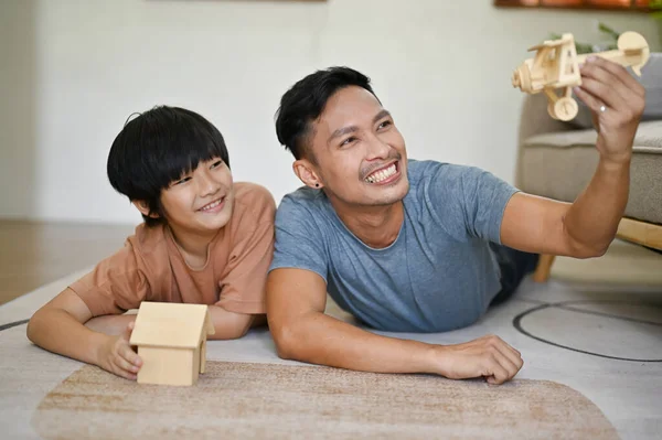 Playful and happy Asian dad playing a wooden airplane model with his son while laying on the living room floor together.
