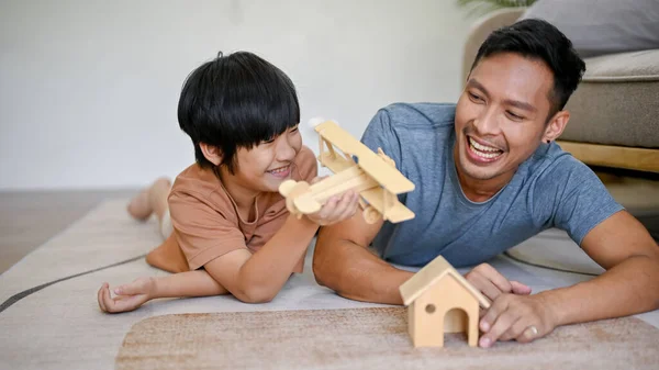 Playful and happy Asian dad playing a wooden figure model with his son while laying on the living room floor together. happy family concept