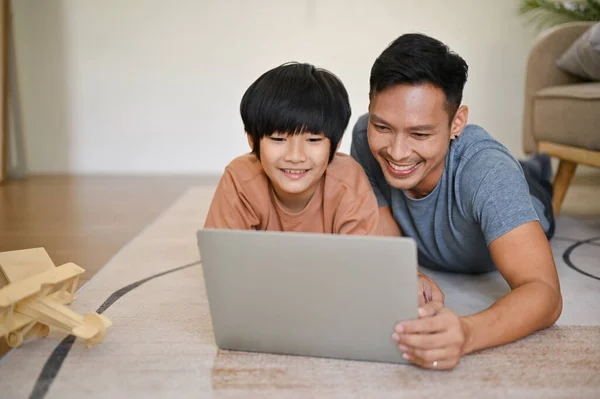 Happy Asian dad is laying on the floor, watching a movie or kid\'s cartoon on laptop, and having fun family time with his son in the living room.
