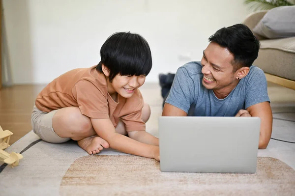 Playful and happy Asian dad is laying on the floor, watching a movie on laptop in the living room with his son on the weekend.