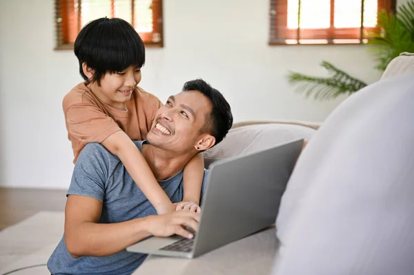 Cute little Asian boy hugging his father from behind while he works on his laptop in the living room. happy family concept