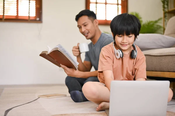 노트북을 사용하는 아시아인 소년은 귀엽지만 아버지는 뒤에서 거실에서 시간을 보낸다 — 스톡 사진