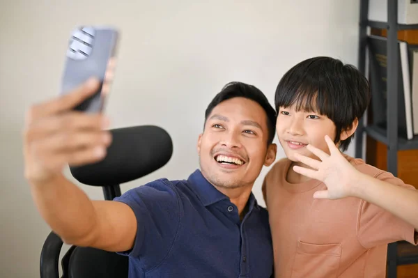 Cheerful and smiling Asian son and dad are taking a picture together with smartphone at home. taking selfie, happy family leisure time