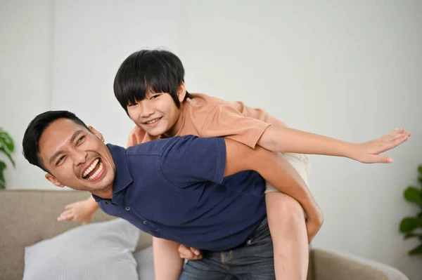 Smiling and laughing Asian dad playing with his son, piggyback, having a fun family time together at home.