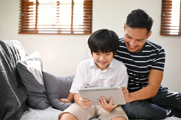 A happy Asian dad enjoys playing online kid\'s games or watching kid\'s video content on a tablet with his son in the living room