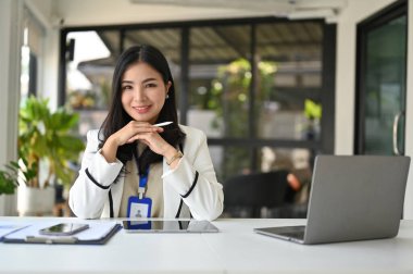 Çekici güzel Asyalı iş kadını ya da bayan pazarlama müdürü masasında elleri çenesinde oturuyor, gülümsüyor ve kameraya bakıyor..