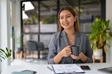 Milenyum Asyalı güzel iş kadını kahvesini yudumluyor, rahatlıyor ve ofis masasında kariyer başarısını hayal ediyor..