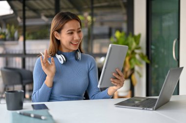 Gülümseyen ve mutlu Milenyum Asyalı kadın serbest kıyafet içinde müşterisiyle birlikte çalıştığı yerdeki bir tabletten video görüşmesi yapıyor..