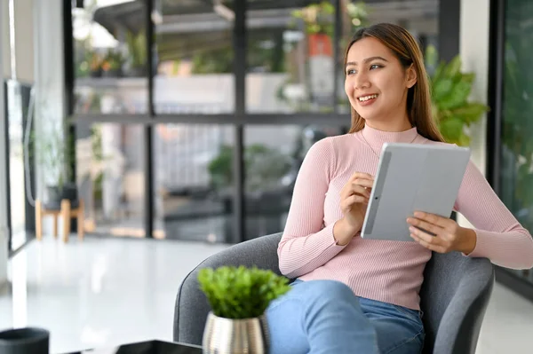 Charming and inspired millennial Asian female freelancer looks out the window for inspiration while using her digital tablet to manage her work while remote working at the cafe.