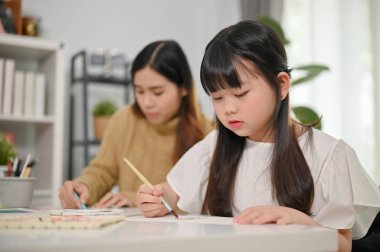 Güzel genç Asyalı kız, annesiyle oturma odasında kağıt üzerinde sulu boyayla resim yapmaya odaklanıyor..