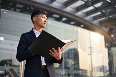 Milenyum Asyalı iş adamı ya da CEO 'su resmi bir iş davasında şirket binasının önünde dururken elinde bir dosya klasörü tutarak ilham verici, kararlı ve kendinden emin bir şekilde duruyor..