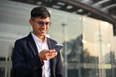 Yakışıklı ve mutlu Asyalı iş adamı veya resmi takım elbiseli erkek CEO şirket binasından çıkarken akıllı telefonunu kullanıyor..