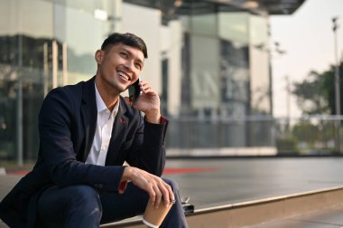Resmi takım elbiseli neşeli Asyalı iş adamı binanın dışında merdivenlerde otururken telefonla konuşuyor. Kentsel yaşam tarzı konsepti