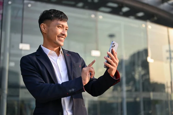 stock image Happy and smiling millennial Asian businessman in a formal business suit using his smartphone while standing outside of the company building.