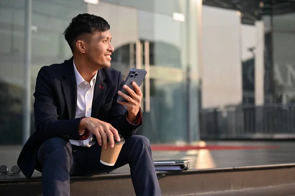 迷人而快乐的千年亚洲商人或穿着正式西装的男工 坐在大楼外的楼梯上 用智能手机喝咖啡 — 图库照片
