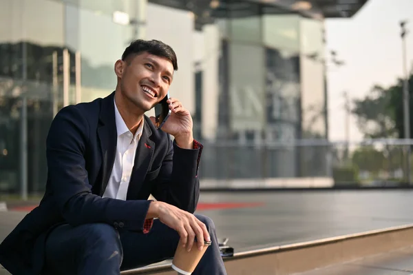 Cheerful millennial Asian businessman in formal business suit talking on the phone with someone while sitting on stairs outside of the building. Urban lifestyle concept