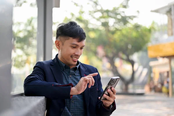 在城市里等公共汽车或出租车时 快乐而英俊的千年亚洲商人用他的智能手机 — 图库照片