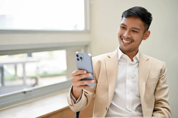 在办公室里放松的时候 穿着正式西装的英俊 微笑的千年亚洲商人用他的智能手机 观看视频 在社交媒体上滚动 — 图库照片