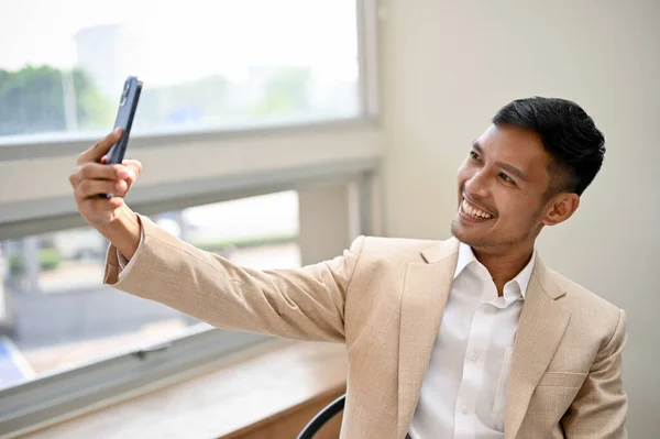 一位英俊 面带微笑的千年亚洲商人坐在办公室里 一边自拍 一边用智能手机录下一段视频 — 图库照片