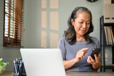 Masasında akıllı telefonunu kullanan mutlu ve çekici bir Asyalı kadın. sohbet, e-posta yanıtlama, mesaj alma, bir web sitesinde kaydırma.