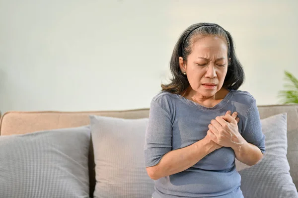 Unwell and sick elderly Asian woman sits on sofa in her living room, touching her chest, suffering from heart attack or chest pain. Health problems concept