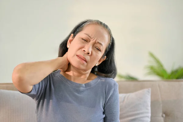 Unhappy senior Asian woman sits on sofa in her living room suffering from neck pain and rubbing her neck.
