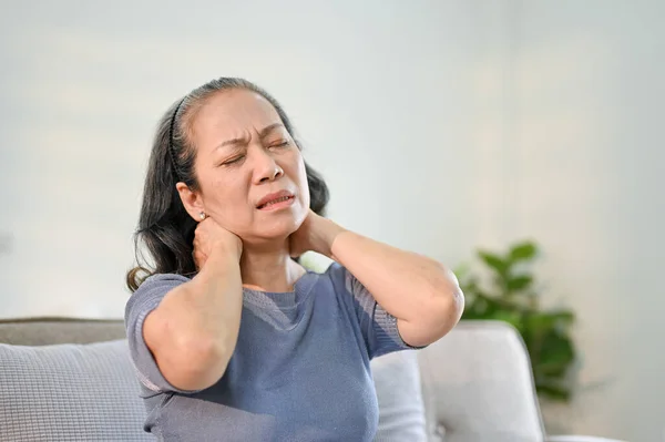 Unhappy elderly Asian woman sits on sofa in her living room suffering from neck pain and rubbing her neck. Health problems concept