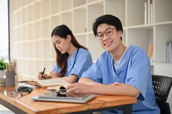 一位英俊聪明的年轻亚洲医学专业学生 穿着制服 和他的朋友在一个图书馆的合作空间里学习 — 图库照片