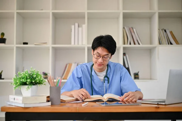 Smart young Asian male medical student in a uniform focuses on reading a book, working on his project, preparing for exam, sitting in the medical school library.