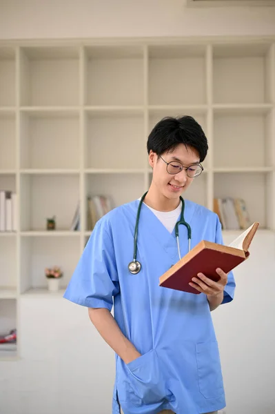 制服を着て眼鏡をかけ 医学校の図書館に立って本を読みながら肖像画 スマート若いアジアの男性医療学生 — ストック写真