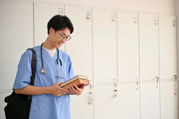 Smart young Asian male medical student in a uniform with medical books and backpack stands in a medical student\'s locker room.