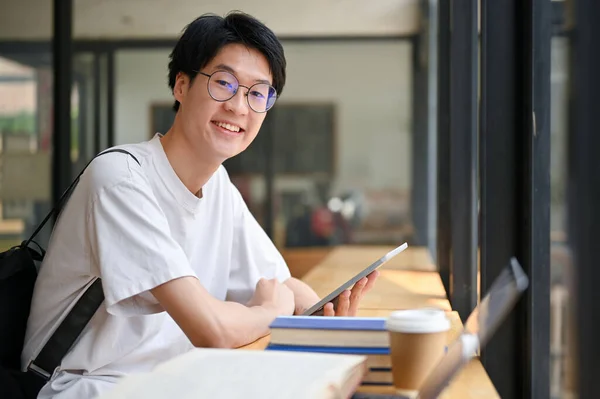 身穿休闲装 面带笑容 英俊的亚裔年轻大学生坐在咖啡店的桌旁 手里拿着平板电脑和课本 — 图库照片