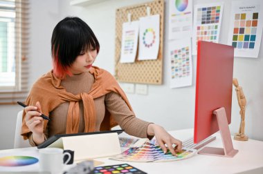 Çekici genç Asyalı kadın web grafik tasarımcısı projesine odaklanıyor, renk örneklerinden renk seçiyor, ofisinde çalışıyor..