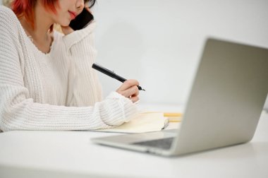 Genç bir Asyalı kadının müşterisiyle telefonda konuşurken not alırken ve ofisinde masasında çalışırken yakın plan görüntüsü..