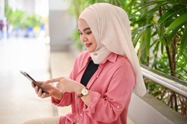 Çekici ve güzel Asyalı genç Müslüman üniversite öğrencisi kampüs binasının koridorunda akıllı telefonunu kullanıyor..