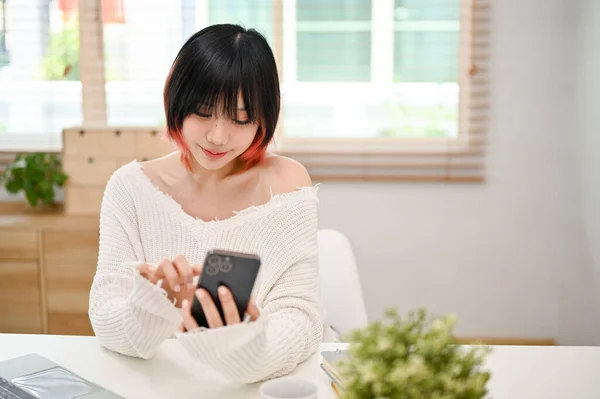 穿着休闲装的年轻亚洲女人很迷人 她在自家的工作空间用智能手机和朋友们聊天 — 图库照片