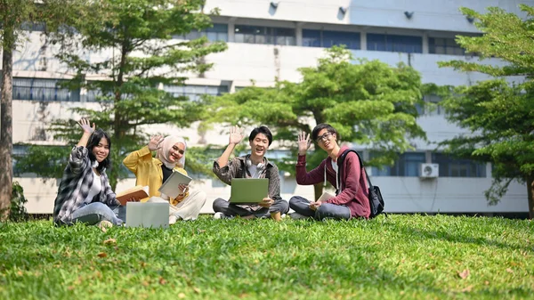一群快乐的来自亚洲的年轻大学生坐在校园公园的草地上 一边挥挥手 一边举手 团队合作 统一的生活方式 — 图库照片