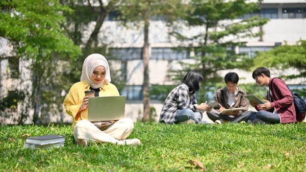 聚焦于年轻的亚洲穆斯林女大学生 穿着头巾坐在草地上 喝着咖啡 在校园公园里用她的笔记本电脑 单一生活方式概念 — 图库照片