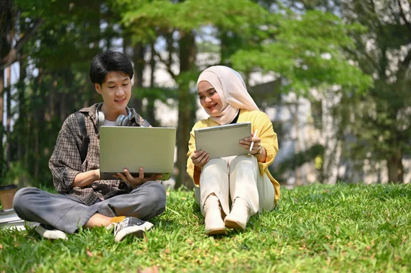 一位年轻英俊的亚洲男性大学生 一边用笔记本电脑 一边带着他的穆斯林女友 一边在校园公园里放松一下 — 图库照片