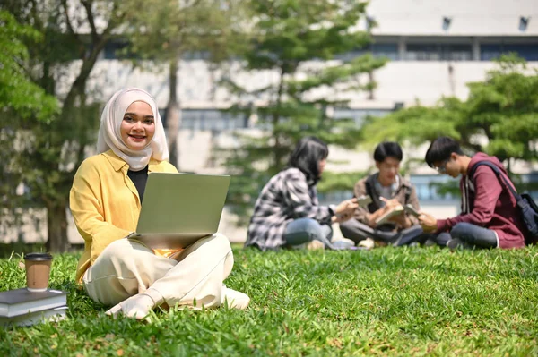 穿着头巾坐在草地上 在校园公园里使用笔记本电脑的年轻的亚洲穆斯林女大学生非常迷人和快乐 — 图库照片